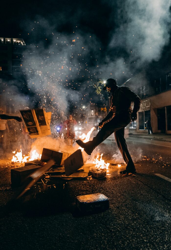 Photograph of the Black Lives Matter protests in Minneapolis. Depicts an individual in front of a fire in the street, with embers and smoke wafting in the air