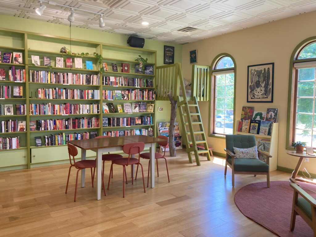 Open space in bookstore, featuring chairs and tables, many books, open windows, and a treehouse in the corner.