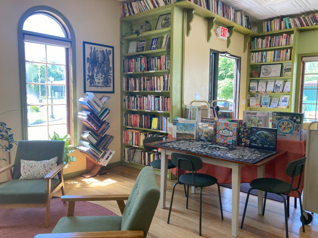 Open space in bookstore, featuring chairs and tables, many books, open windows, the puzzles table and puzzles for sale.