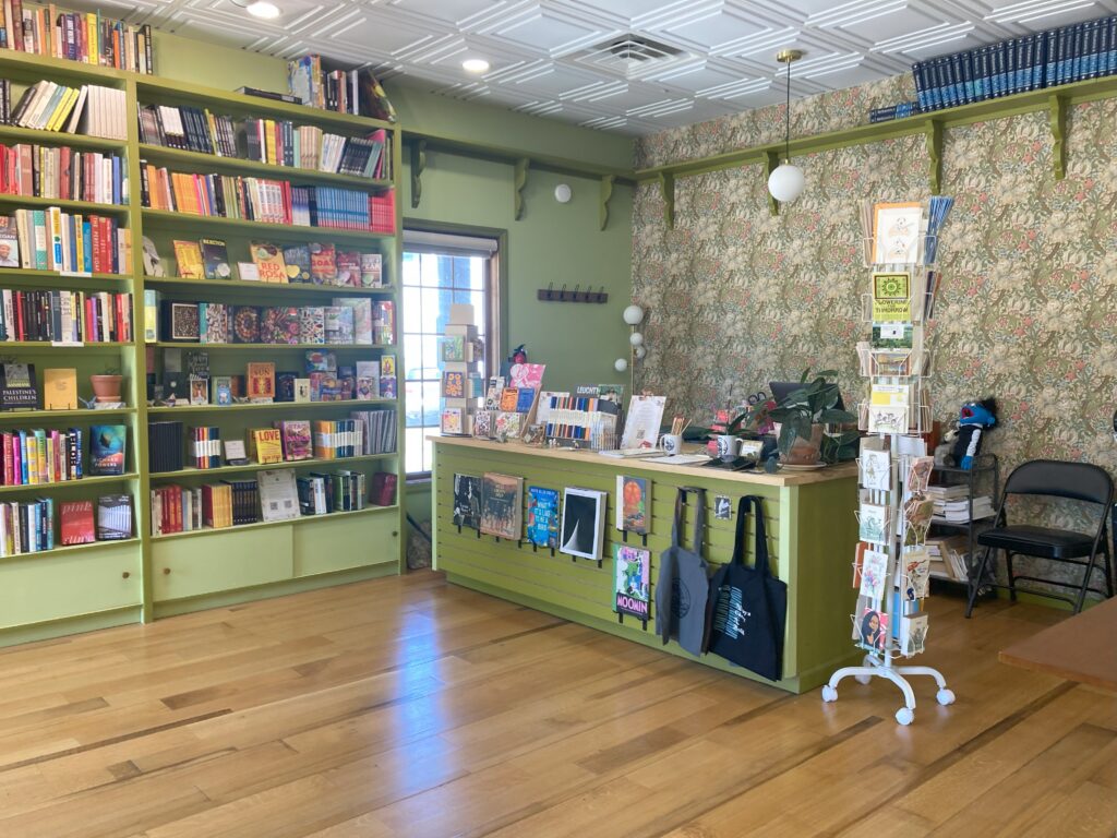 Front desk of store, filled with books, greeting cards, notebooks and journals, and so on. Features colorful William Morris wallpaper on the backwall