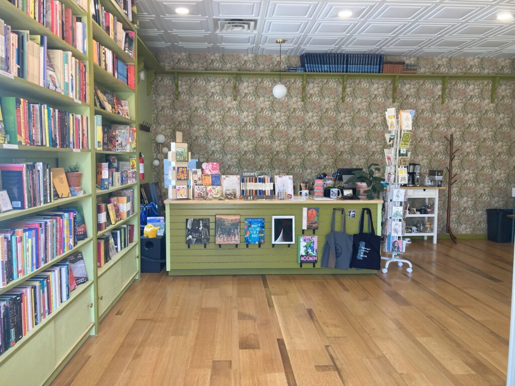 Front desk of store, filled with books, greeting cards, notebooks and journals, and so on. Features colorful William Morris wallpaper on the backwall
