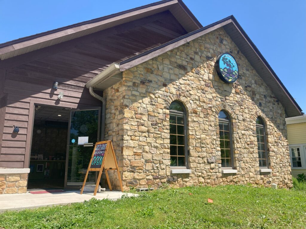 Exterior of Redbud Books. A warm, sandstone facade with three large windows, the Redbud logo in color on a sign near the roof, and open door, and a sandwich board sign that reads "Redbud Books, Open"